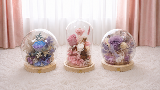 Three preserved flower domes with glass covers on a white carpet — blue, pink, and lavender arrangements from Fu Bloom Flowers Singapore